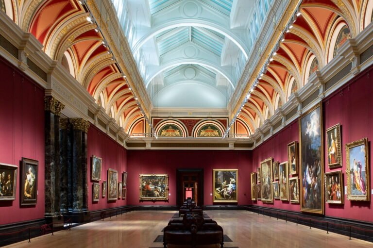 Grand gallery interior with classical paintings displayed on crimson walls beneath an ornate vaulted ceiling
